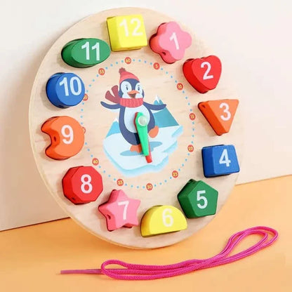 Ceas Montessori din lemn cu pinguin, forme colorate și șnur roz pe fundal cald.