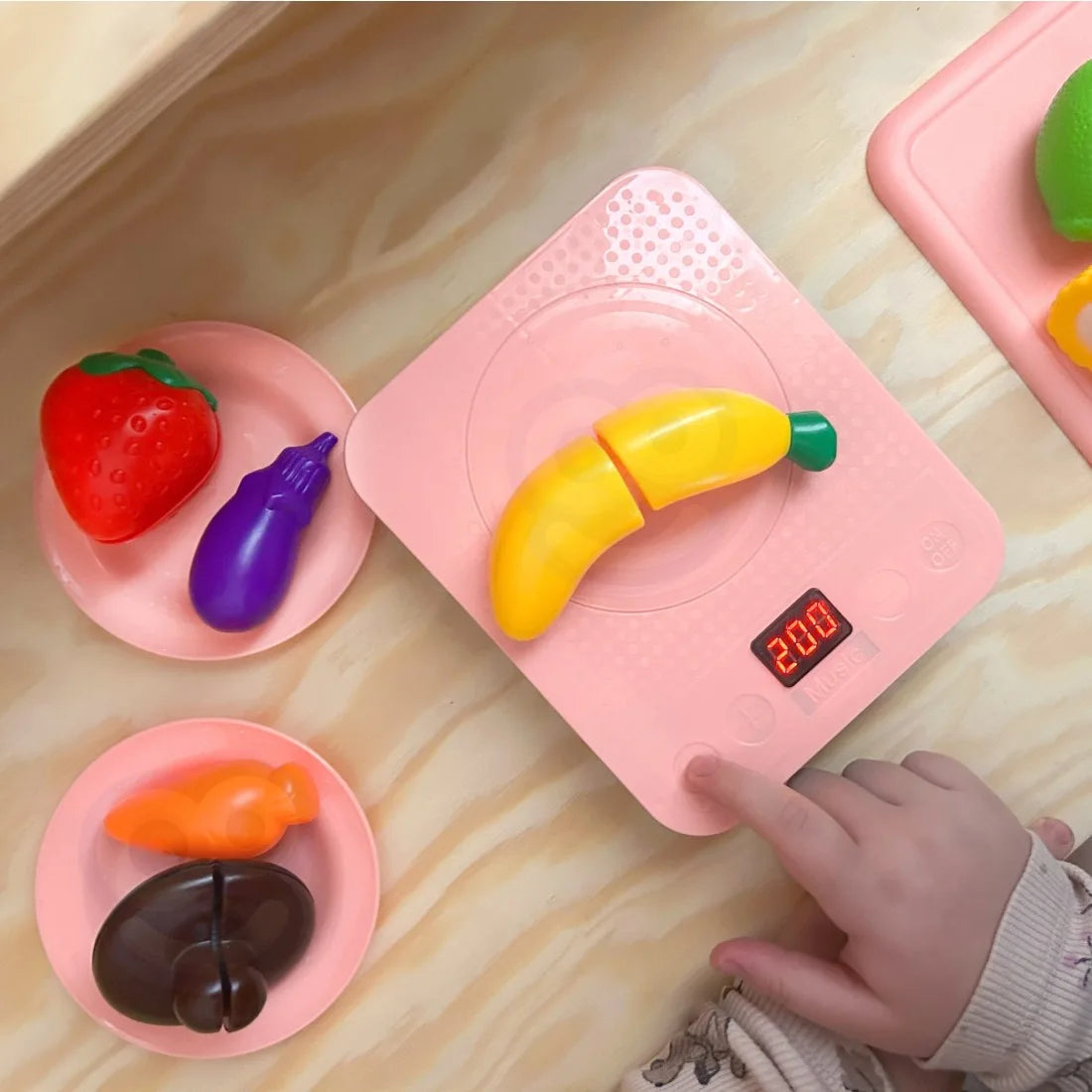 Copil care cântărește legume de jucărie pe plita roz din setul Woopie Magic Kitchen Sink.