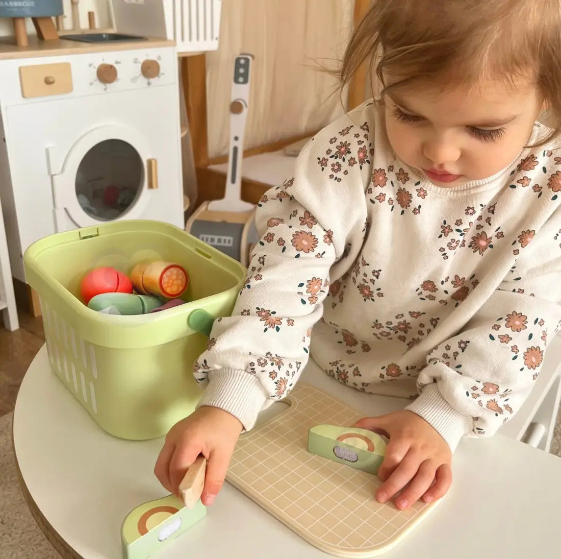 Copil care taie avocado din lemn Tooky Toy pe tocător, cu coșul de alimente aproape pentru joc de rol.