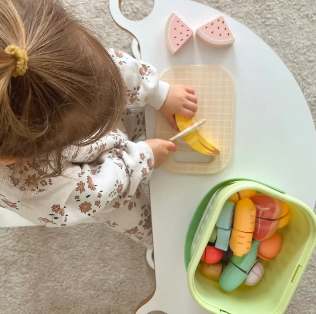 Copil văzut de sus în timp ce taie banana din lemn Tooky Toy pe tocător, lângă coșul cu alimente.
