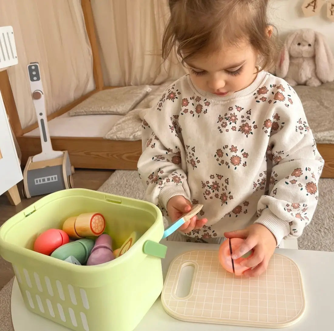 Copil care taie o roșie din lemn Tooky Toy cu cuțitul, pe tocător, lângă coșul verde de depozitare.