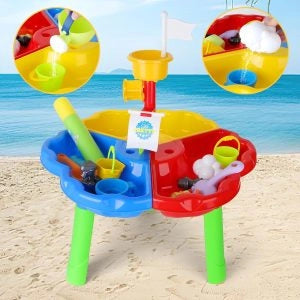 Masă Woopie Sand and Water Play Table pe plajă, cu găletușe, turn de apă și compartimente pentru nisip.